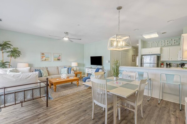 Un salon moderne avec un canapé, une table basse et un coin repas. On y trouve également un téléviseur, un ventilateur de plafond et une cuisine avec des meubles blancs.