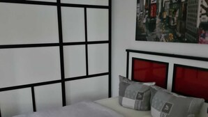 Un dormitorio con cama, cabecera decorada con una pintura de paisaje urbano y ventana con vista a edificios.