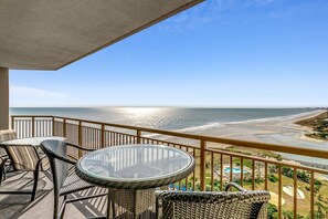 A balcony with wicker furniture and a glass table overlooking a beach and ocean.