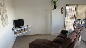 Una sala de estar con un sofá marrón, un televisor de pantalla plana sobre un estante blanco, una planta en maceta y un comedor con mesa y sillas.