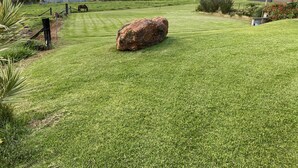 Zona verde bien cuidada con una roca grande, una valla y un edificio al fondo.