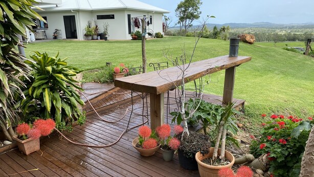 Una terraza de madera con plantas en macetas y una mesa, con vista a un jardín de césped y una casa blanca.