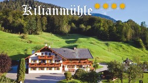 A large, traditional chalet with a sloped roof, surrounded by lush green hills and a clear sky.