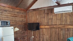 Une pièce rustique avec des murs en bois, un ventilateur de plafond, un téléviseur fixé au mur, un réfrigérateur blanc et une chaise bleue.