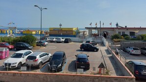 Ein Parkplatz mit mehreren Autos, ein Gebäude mit gelber Fassade und im Hintergrund freie Sicht auf das Meer.