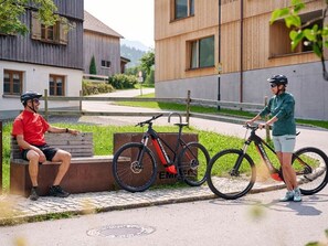 Dos personas sentadas y de pie cerca de bicicletas en un edificio moderno con zona pavimentada y vegetación.