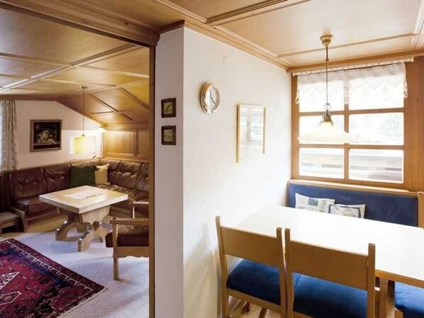 A room with a dining table, chairs, a sofa, a window with curtains, and a clock on the wall.