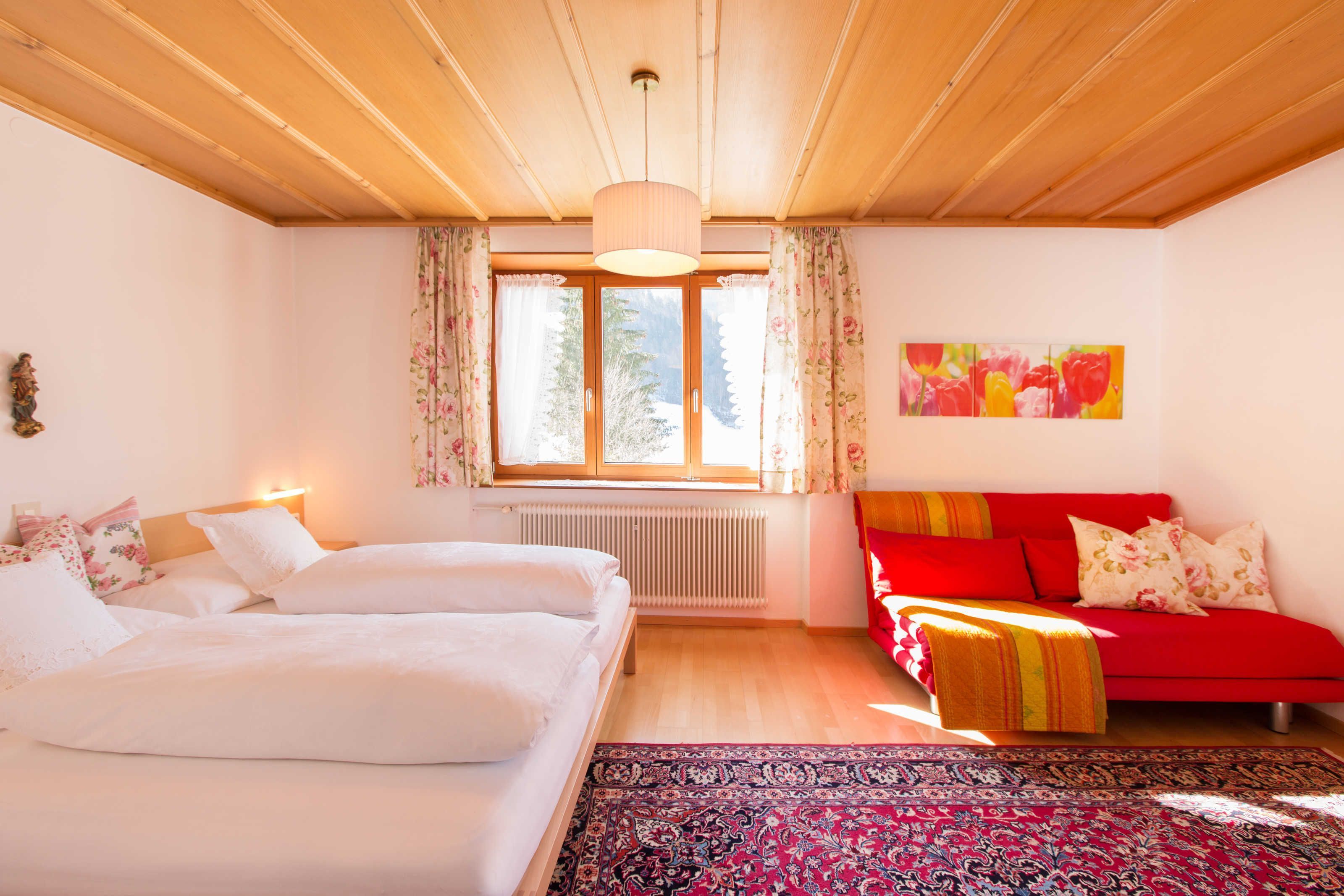 Une chambre à coucher avec un lit, un canapé rouge, une fenêtre avec des rideaux, un radiateur et un tableau accroché au mur.