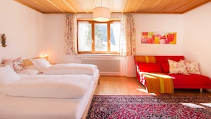 Un dormitorio con cama, sofá rojo, ventana con cortinas, radiador y un cuadro en la pared.