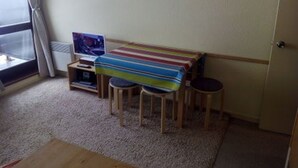 A small, cozy room with a bed, a TV, and a colorful striped blanket. There are two wooden stools and a small table with a remote control.