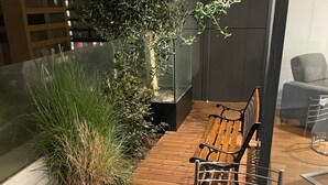 Een moderne buitenruimte met een houten bank en een ronde tafel, omgeven door een glazen constructie en weelderige beplanting.