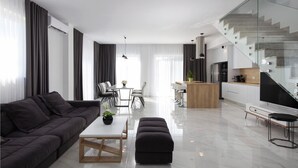 A modern living room with a dark sofa, a white coffee table, and a dining area with a glass table and chairs. There is a kitchen with white cabinets and a stainless steel refrigerator. A glass staircase leads to an upper level.