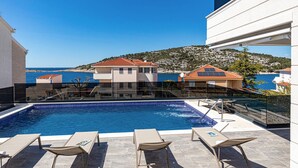 A swimming pool with lounge chairs, a clear view of the sea, and buildings with red-tiled roofs.