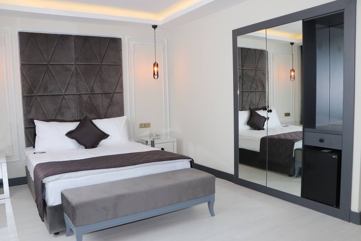 A modern bedroom with a large bed, a bench, a nightstand, a mirror, and a wardrobe.