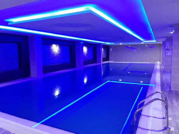 A modern indoor swimming pool with blue lighting, clear water, and a metal ladder.
