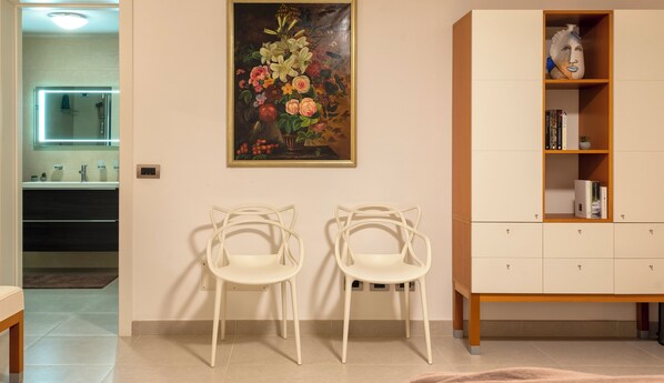 A room with two white chairs, a painting of flowers, a wooden cabinet with books, and a bathroom visible in the background. - Alghero Spectacular sea view, for exclusive use, villa with private pool (Alghero)