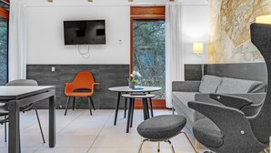A modern living room with a grey sofa, orange chair, and a small round table with a plant. A flat-screen TV is mounted on the wall, and there's a window with a view of trees. - Awesome home in Grenaa with WiFi (Grenaa)