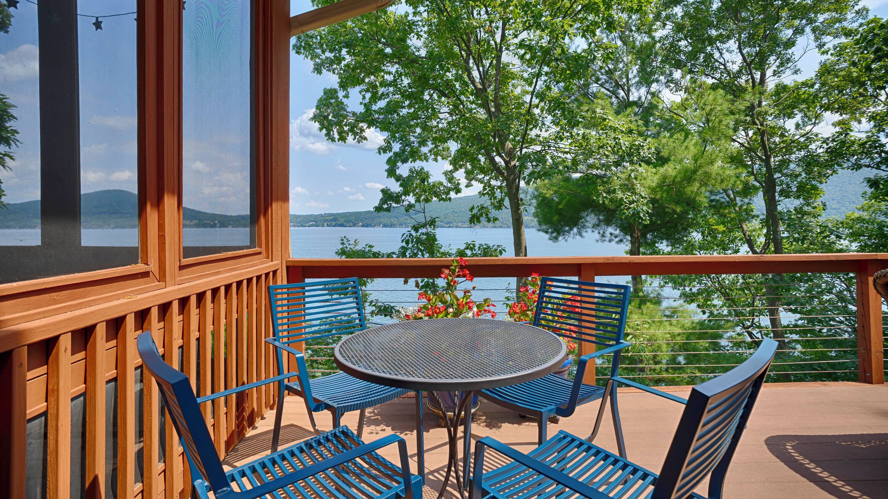 Un balcon avec vue sur un lac et des arbres, agrémenté de chaises en métal bleues et d’une table ronde.