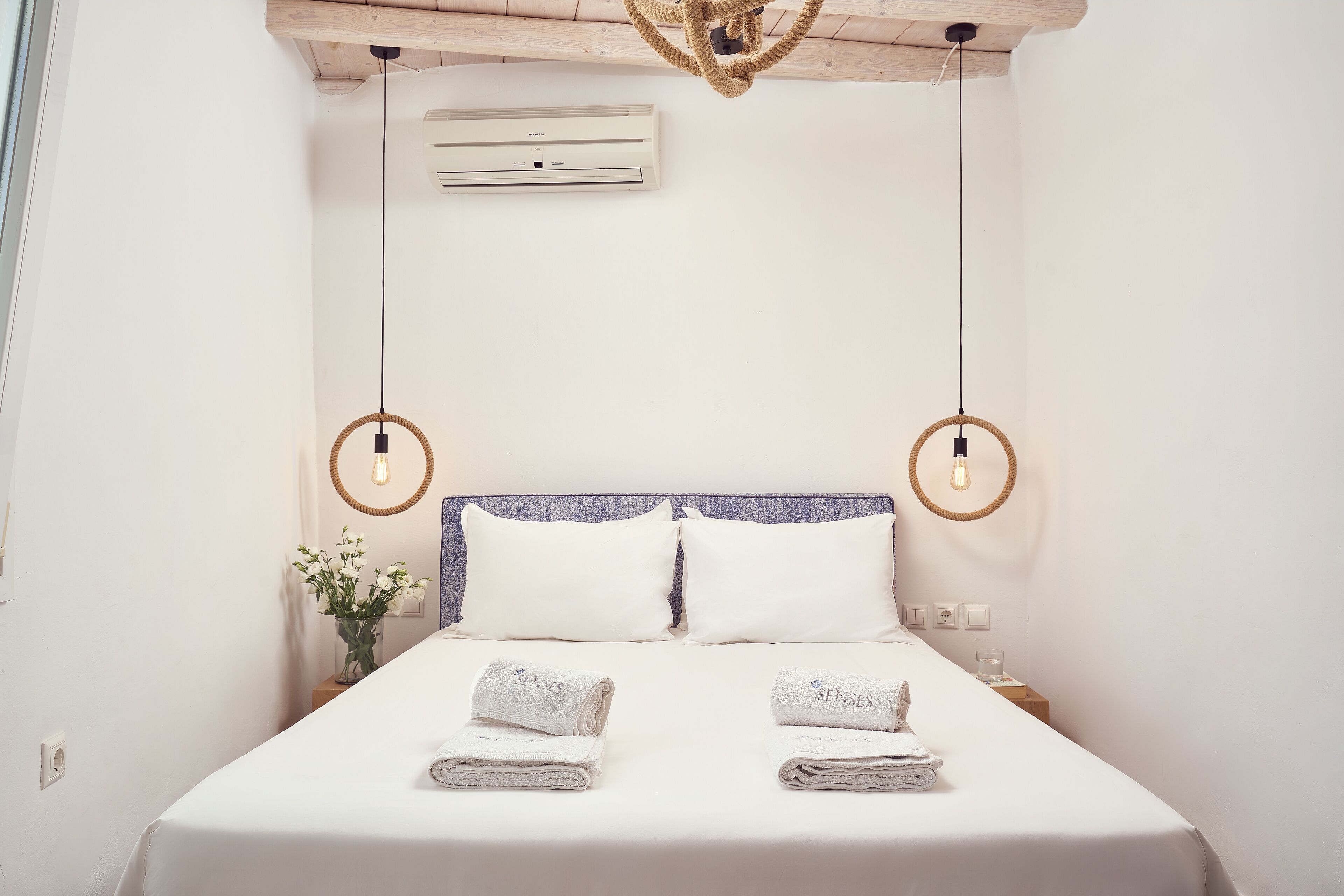 Un letto rifatto con lenzuola bianche e due asciugamani con la scritta 'SENSES', un soffitto in legno, un condizionatore d'aria a parete e un piccolo vaso con fiori bianchi sul comodino.