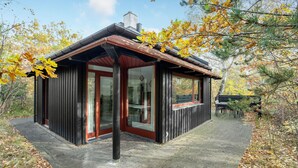 Une petite cabane avec une porte rouge et des fenêtres, entourée d’arbres aux feuilles d’automne.