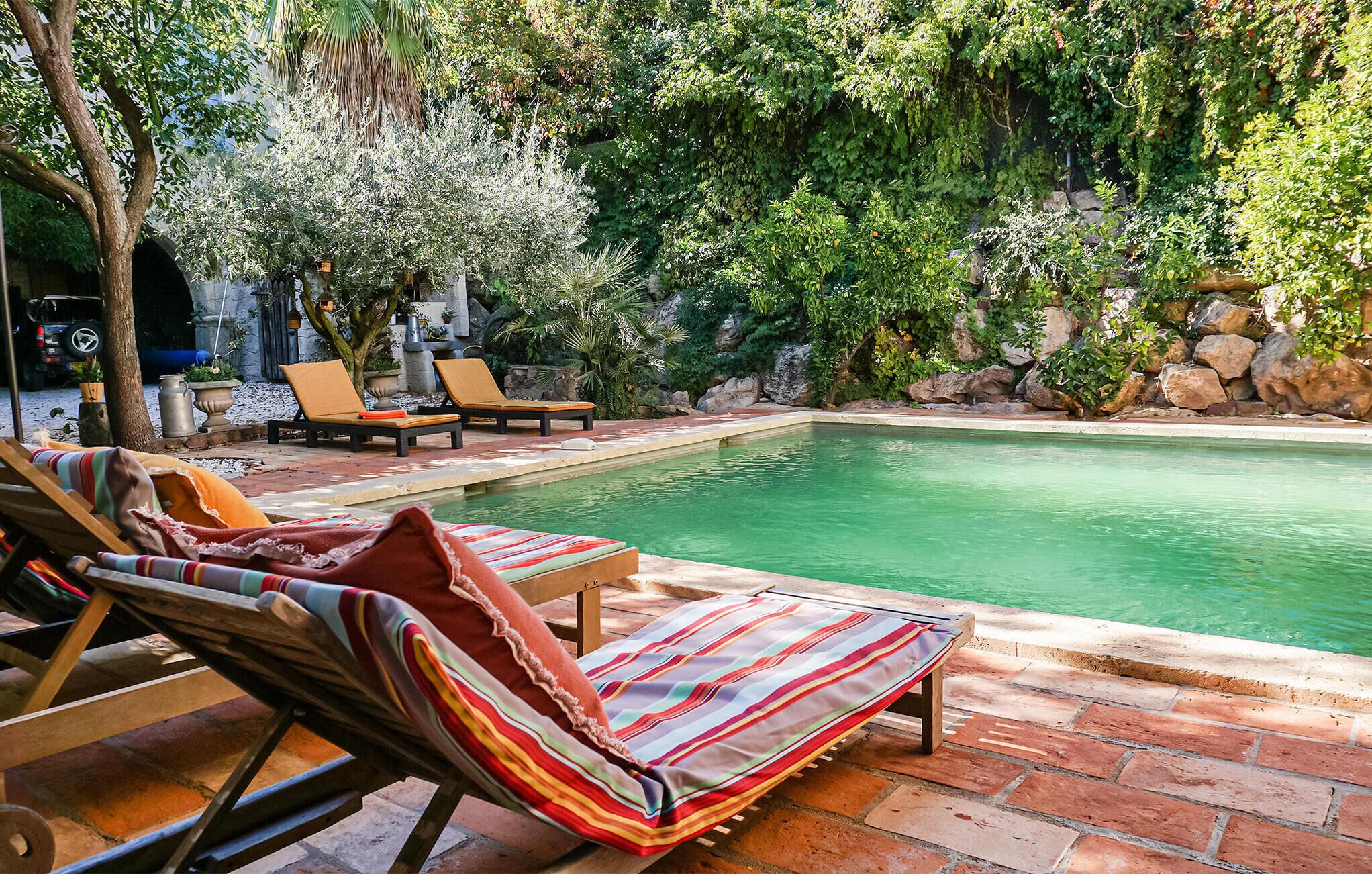 Un área de piscina con sillones reclinables y una pequeña mesa, rodeada de exuberante vegetación y rocas.