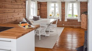 A cozy cabin-style room with a wooden interior, a dining table with chairs, a kitchen area with a sink and a fruit plate, and large windows with curtains. - 3 bedroom cozy home in Lindesnes (Lindesnes)