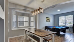 A pool table in a room with a wooden table and bench, large windows with blinds, and a ceiling light fixture.