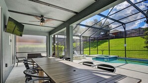 A covered outdoor area with a long table and chairs, a ceiling fan, a TV screen, and a view of a swimming pool and green lawn.