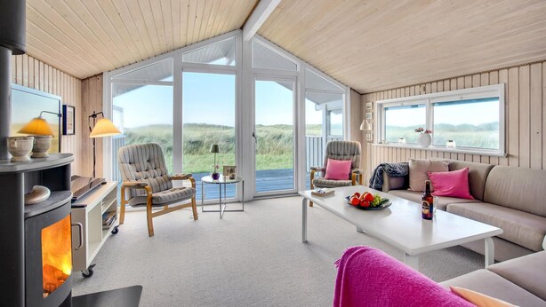 Una habitación acogedora con chimenea, dos sillones, un sofá y una mesa de centro. Amplios ventanales ofrecen vistas al exterior.