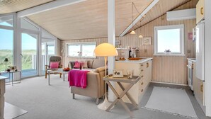 Amplia sala de estar con sofá y sillones, una mesa de comedor y una cocina con paneles de madera y electrodomésticos blancos.
