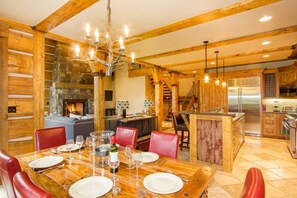 Um interior rústico com uma mesa de jantar de madeira posta para seis pessoas, uma lareira e uma área de cozinha com eletrodomésticos de aço inoxidável.