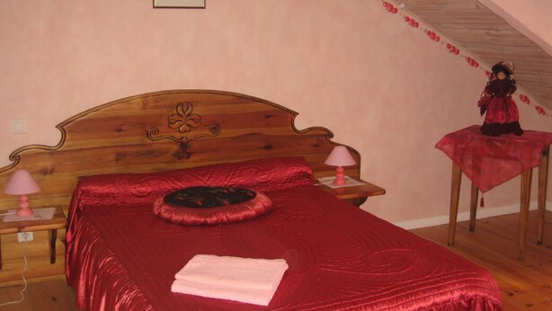 Un dormitorio con una cama de madera, una colcha roja, una toalla rosa, una mesita de noche con una lámpara y una pequeña mesa con una muñeca.
