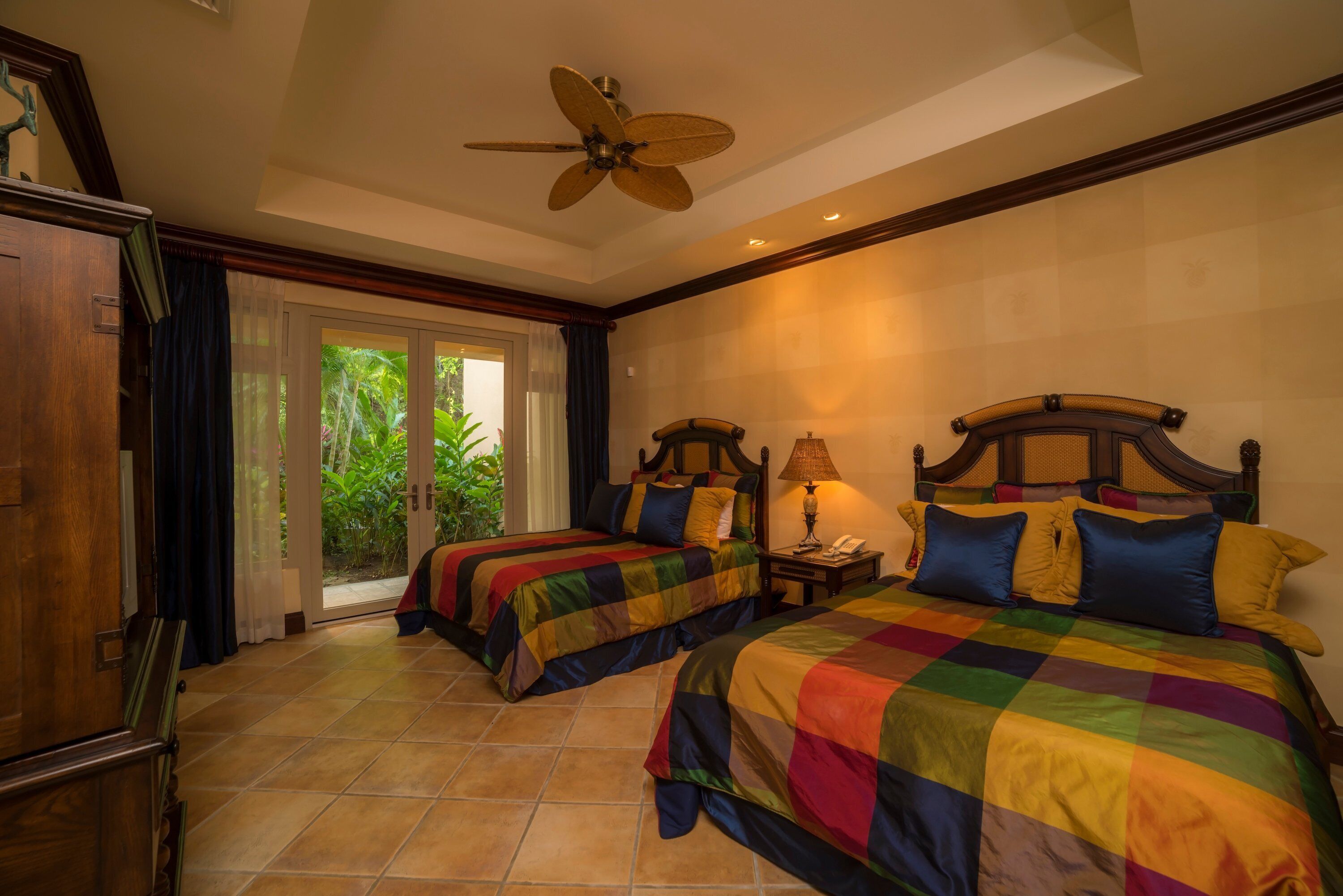A hotel room with two beds, a ceiling fan, a nightstand with a lamp, and a view of greenery through the sliding glass door.