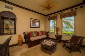 A living room with a sofa, a coffee table, a chair, a large fan, a window with a view, and a decorative vase.