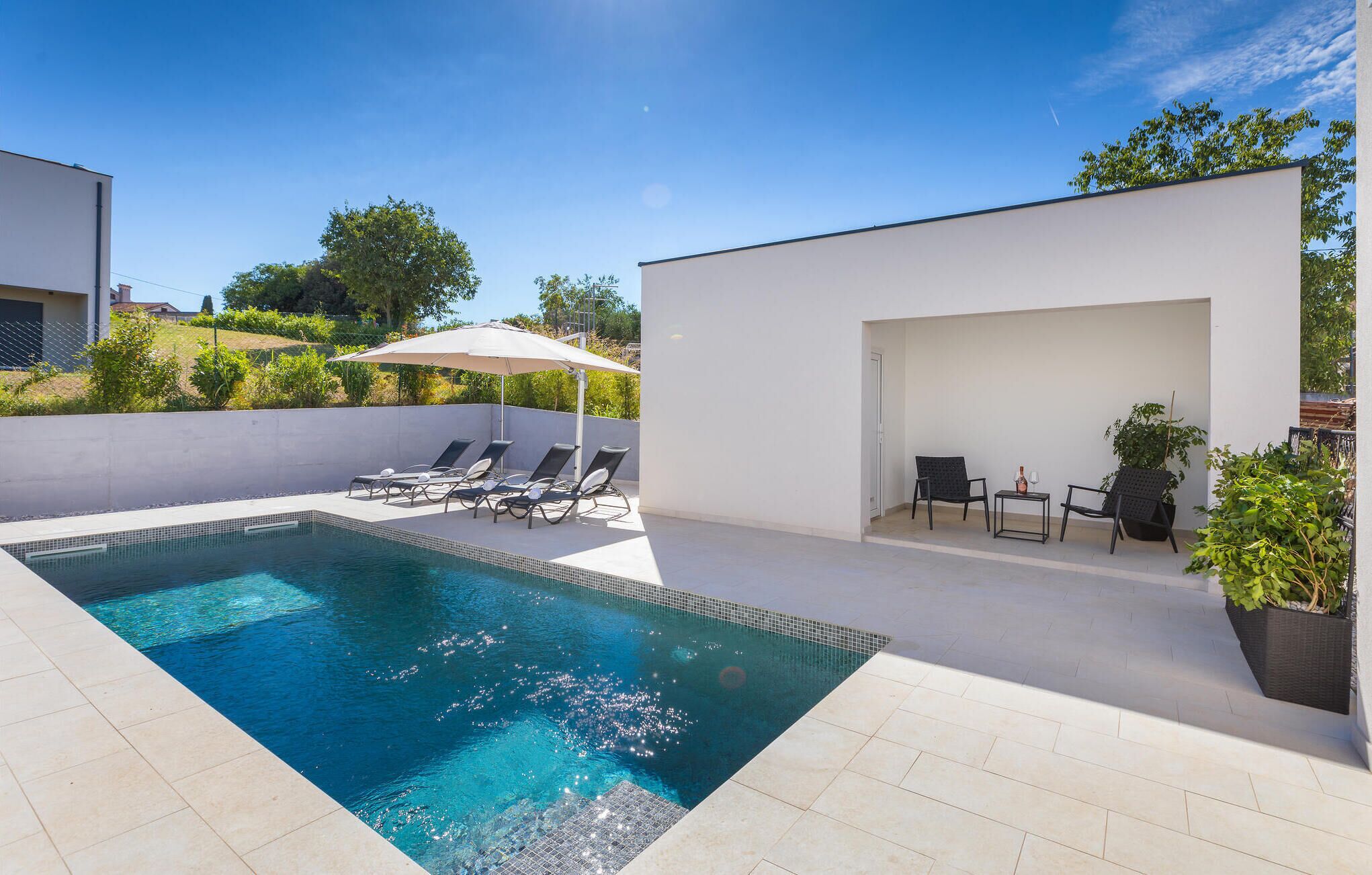 A modern outdoor pool area with a clear blue pool, sun loungers, a covered seating area, and potted plants.