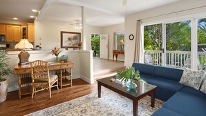 Un vaste espace de vie comprenant un canapé bleu, une table basse en bois et une table à manger avec des chaises. On aperçoit un ventilateur de plafond et on peut admirer la vue d’un balcon aménagé avec du mobilier d’extérieur.