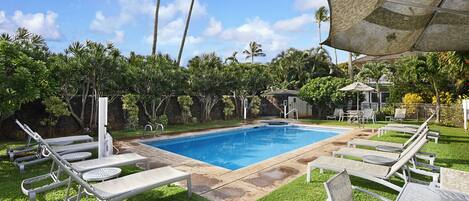 Una piscina con sillas de descanso y una zona cubierta, rodeada de follaje y un cielo despejado.