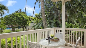 Une terrasse blanche avec une table et des chaises en verre, entourée d’une végétation tropicale luxuriante et offrant une vue sur une piscine.
