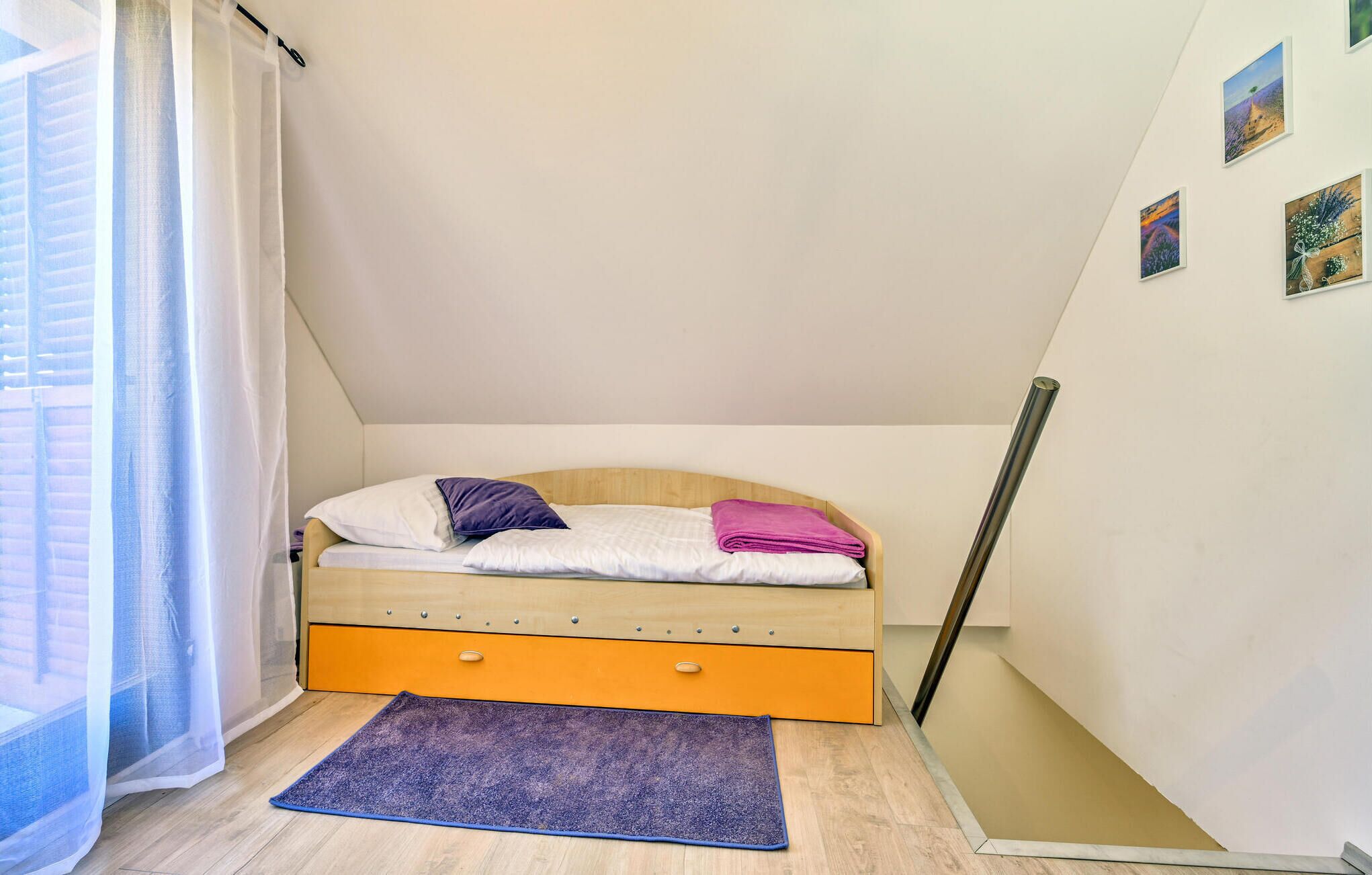 Een kleine eenpersoonskamer met een houten hoofdbord, een paarse plaid en een blauw vloerkleed.