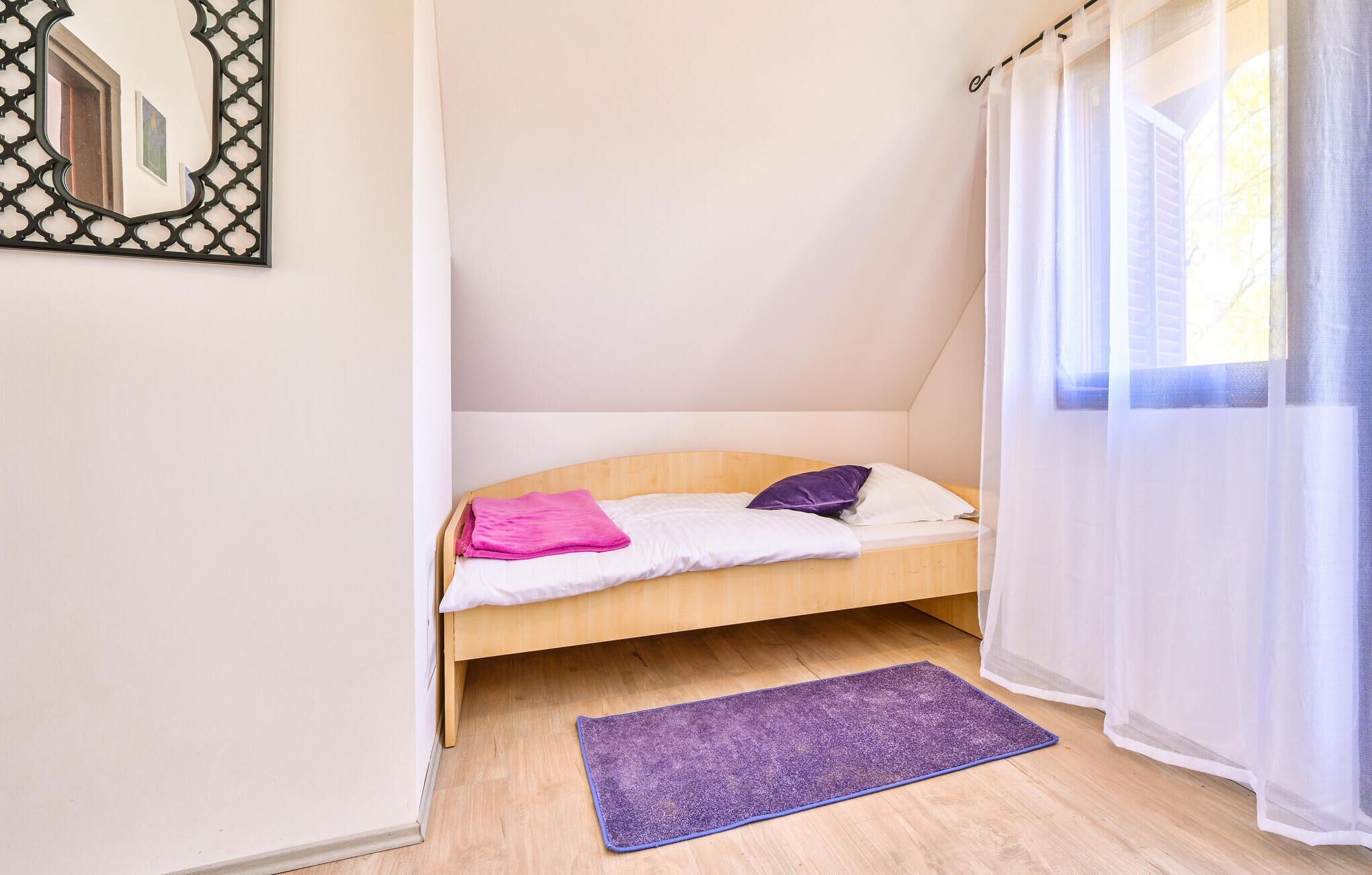 Une petite chambre avec un lit simple, une tête de lit en bois, un tapis violet et une fenêtre ornée de rideaux légers.