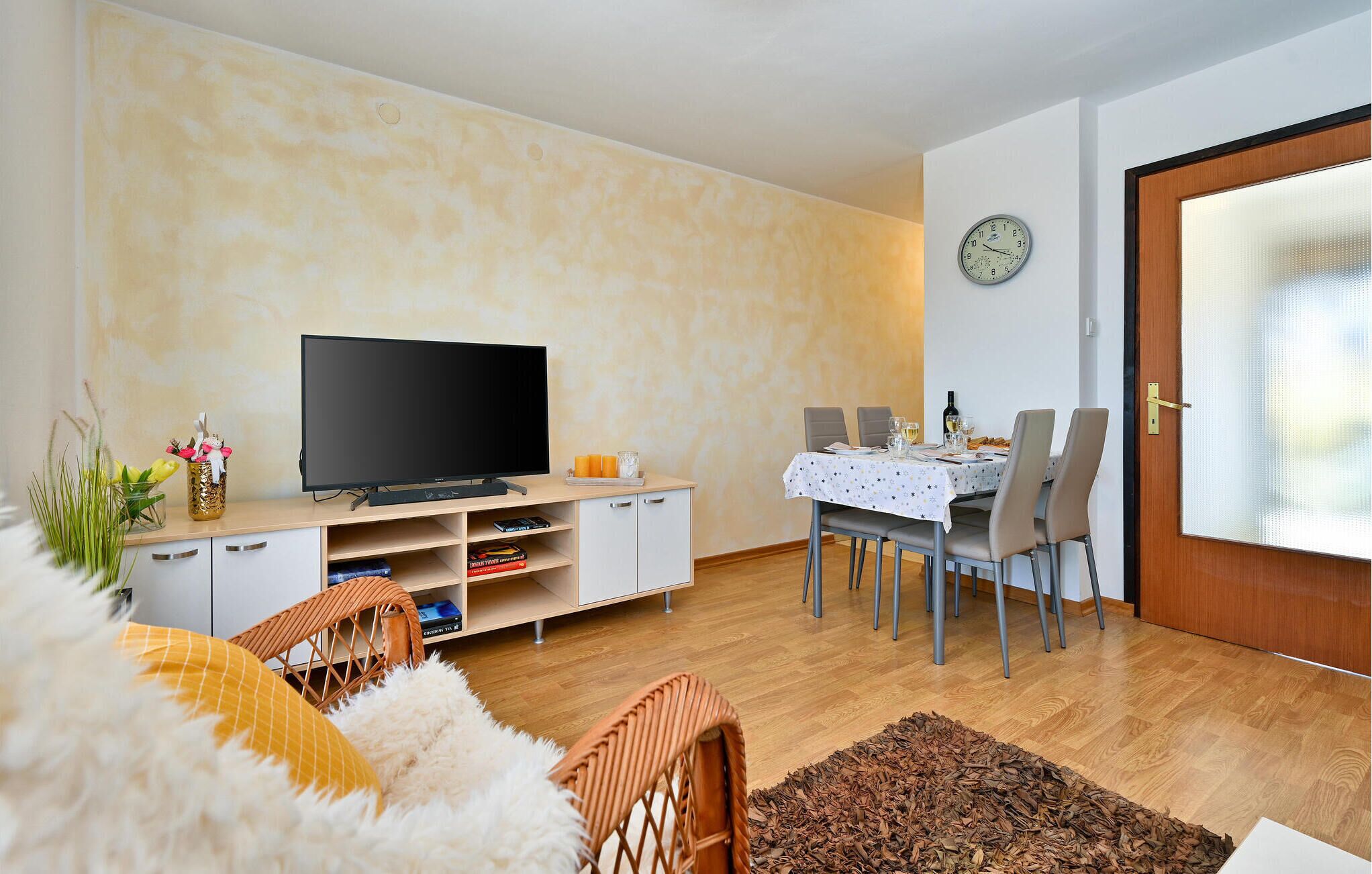 Un salon avec un téléviseur à écran plat, une table à manger dressée pour deux personnes et une horloge au mur.