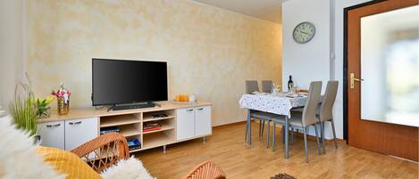 Un salon avec un téléviseur à écran plat, une table à manger dressée pour deux personnes et une horloge au mur.