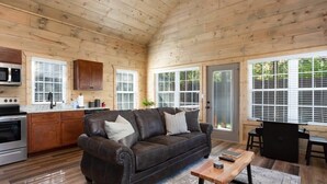 A cozy cabin interior with a wooden ceiling, a kitchen area with a stove and microwave, a leather sofa with cushions, a wooden coffee table, and large windows with blinds.