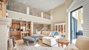 A spacious living area with a high ceiling, a staircase to an upper level, a kitchen with wooden cabinets, and a large window providing natural light.