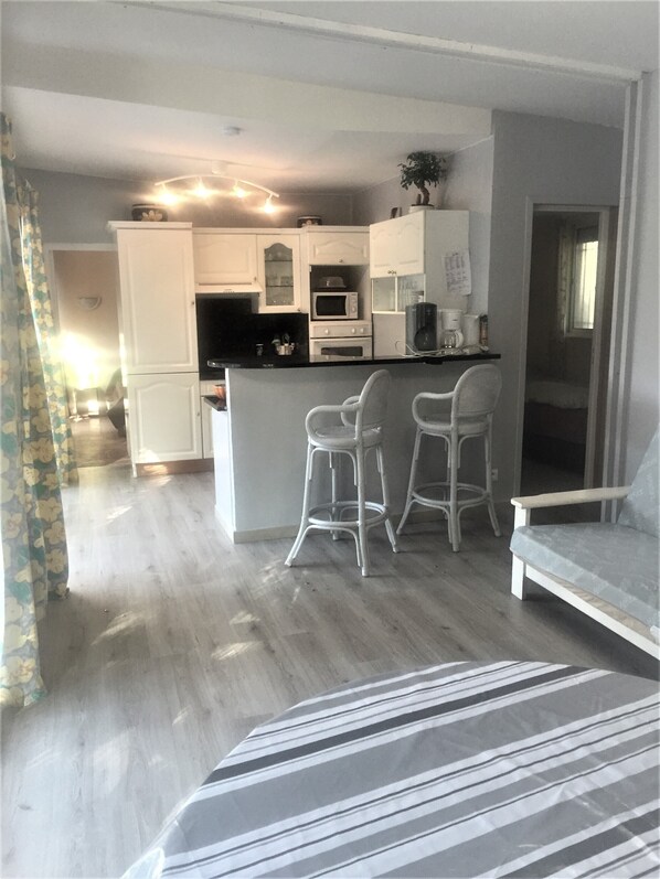 A compact kitchen with white cabinets, a microwave, and a coffee maker. There are two white bar stools and a white sofa in the living area.