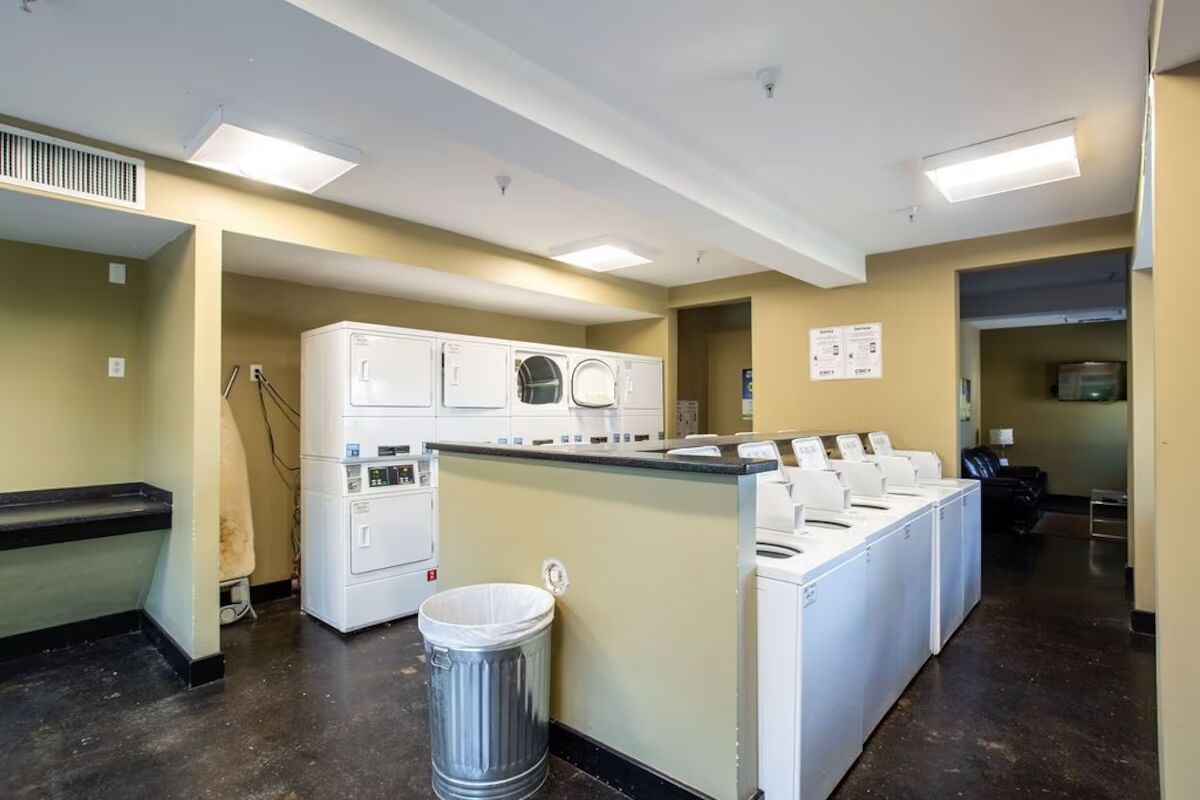 A laundry facility with multiple washing machines and dryers, a trash can, and a bulletin board on the wall.