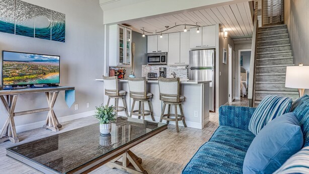 A modern living room with a glass coffee table, a blue sofa, and a TV on a wooden stand. - Great location, Stylish Beachy Condo With Upgraded Amenities & Ocean View (Kihei)