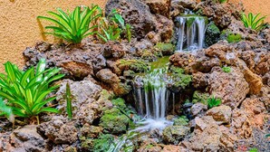 A small, rocky garden with two waterfalls and green plants. - Great location, Stylish Beachy Condo With Upgraded Amenities & Ocean View (Kihei)