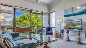 A modern living room with a glass coffee table, blue sofa, and a television on a wooden stand. - Great location, Stylish Beachy Condo With Upgraded Amenities & Ocean View (Kihei)