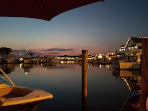 A marina at dusk with boats docked, a wooden post in the foreground, and a building with lights in the background.